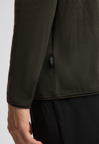 Donkergroen overhemd met lange mouwen van een textuurstof, zichtbare stiksels en een klein logotagje aan de zijkant. Gecombineerd met zwarte broek.