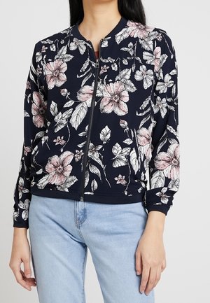 Giacca a zip blu navy con stampa floreale rosa e bianca, indossata con jeans azzurri.