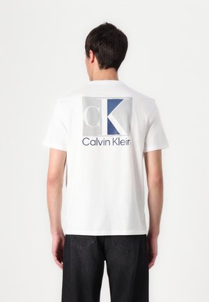 Mężczyzna w białej koszulce z dużym logo Calvin Klein w kolorze niebieskim i szarym na plecach, w czarnych dżinsach, stojący tyłem.