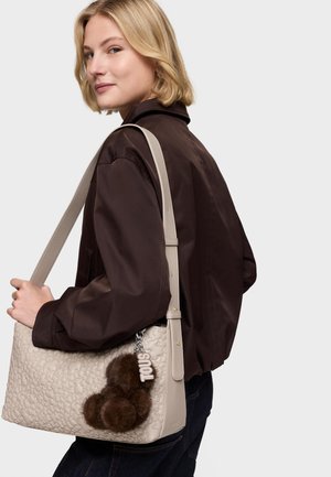 Mujer rubia con chaqueta marrón oscuro que lleva un bolso beige texturizado con pompones de piel marrón y un llavero "TOUS" colgado del hombro.