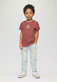 Bruine katoenen T-shirt met multikleurige grafische ontwerpen, gecombineerd met lichtblauwe jeans en witte sneakers met grijze accenten.