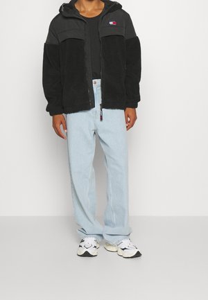 Osoba ubrana w czarną polarową kurtkę z kapturem z logo flagi, jasnoniebieskie dżinsy i białe trampki stojąca na białym tle.