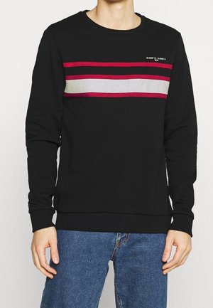 Homme portant un pull noir avec des rayures horizontales rouges et grises sur la poitrine, associé à un jean bleu, sur un fond uni.