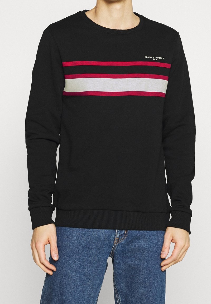 Homme portant un pull noir avec des rayures horizontales rouges et grises sur la poitrine, associé à un jean bleu, sur un fond uni.