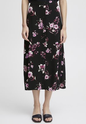 Kvinde iført en sort midi nederdel med knapper foran med pink og hvide blomsterprint og sorte åbne tøfler.
