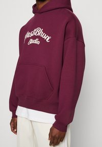 Person i en vinröd hoodie med texten "Mostbhvn Studio", vit skjorta under och ljusbeiga byxor, som står framför en enkel bakgrund.