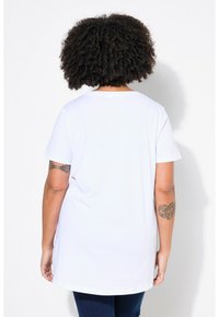 Camiseta blanca de manga corta y escote redondo. Tejido suave, corte relajado y dobladillo trasero más largo. Tatuajes visibles en los brazos.