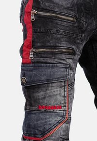 Zwarte denim cargobroek met rode accenten en meerdere ritszakken. Kenmerkend zijn de textuurstof en de geborduurde logo-detail.