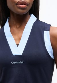 Γυναίκα που φοράει αμάνικη μπλε μπλούζα με φωτεινό γαλάζιο γύρω στο λαιμό και το λογότυπο «Calvin Klein» στο κέντρο κάτω από το λαιμό.
