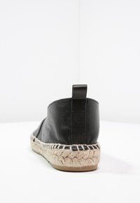 Zapato de cuero negro con suela de yute de espadrille, forma redondeada, parte superior texturizada y tirador trasero. La suela presenta un diseño trenzado.