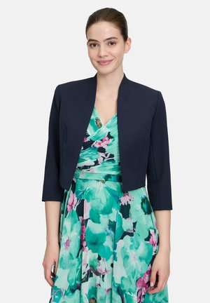 Giacca cropped blu navy con taglio dritto, maniche 3/4 e design minimalista, indossata sopra un vestito floreale teal e rosa con scollatura a V.