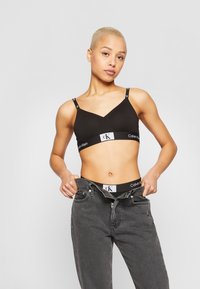 Kvinne med kort blondt hår iført svart Calvin Klein bralette og truser, som drar ned grå jeans mot en grå bakgrunn.