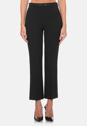 Donna che indossa pantaloni neri attillati e corti con una cintura sottile e scarpe nere con tacchi alti a punta, in piedi davanti a uno sfondo semplice.