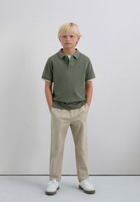 Camisa polo verde de manga corta, con cuello y detalles en blanco, combinada con pantalones beige y zapatillas blancas. Diseño sencillo.