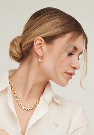 Femme blonde avec les cheveux en chignon portant un chemisier blanc, un collier chaîne en or et des boucles d'oreilles créoles assorties en or, regardant vers le bas et sur le côté.