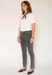 T-shirt blanc, jean gris slim-fit, et chaussures noires à enfiler avec des accents décoratifs. Écharpe marron nouée au cou pour ajouter du détail.
