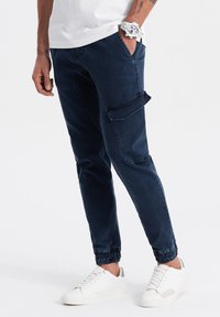 Pantalones jogger azul marino con puños elásticos y un bolsillo tipo cargo. Hechos de algodón, con una textura suave y un corte ajustado. Combinados con zapatillas blancas.