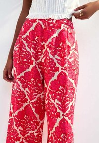 Weite Hose mit einem lebhaften pinken Blumenmuster auf einem cremefarbenen Hintergrund, aus leichtem Stoff mit glatter Textur.