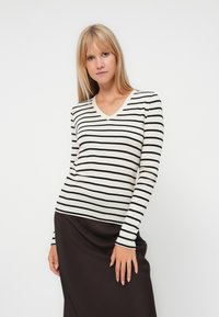Crème kleurige V-hals top met lange mouwen en zwarte horizontale strepen; aangesloten ontwerp, zachte textuur, gedragen met een donkere rok, minimale accessoires.