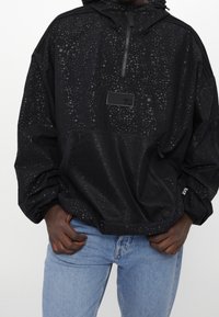 Man i svart mönstrad huvtröja med dragkedja på bröstfickan och nätficka på framtiden, tillsammans med ljusblå jeans.