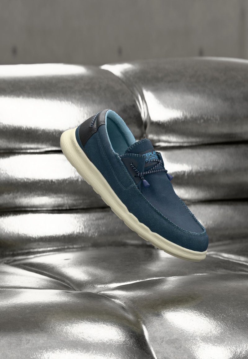 Blauer Slipper mit weißer Sohle und schwarzem Rand, der über einer glänzenden, metallisch-silbernen, gesteppte Oberfläche schwebt.