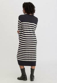 Gestreiftes, marineblaues und cremefarbenes Strickkleid mit langen Ärmeln, gerippten Bündchen und einem knielangen Saum. Das Model trägt schwarze Lederstiefel bis zur Knöchel.