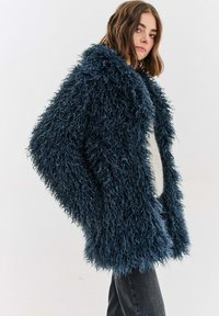 Bunda z umelého kožušiny v tmavomodrej farbe s textúrovaným, kožušinovým povrchom. Má široký golier a otvorený predný dizajn, nosená cez ľahký sveter.