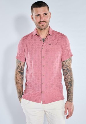 Mann mit kurzen Haaren und Tattoos trägt ein rosa gemustertes kurzärmeliges Hemd und weiße Hose, stehend vor einem schlichten Hintergrund.