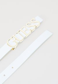 Hvit lærbelte med en gullfarget metallspenne der ordet "GUESS" er tydelig vist. Glatt tekstur med hull for justering.