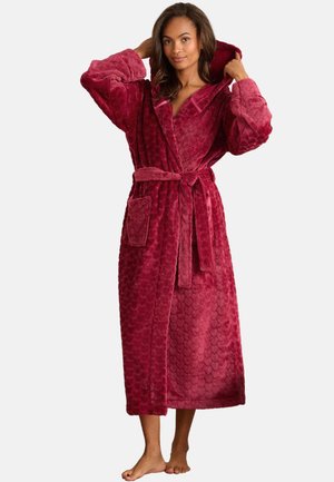 Donna che sistema il cappuccio di un lungo accappatoio in peluche rosso scuro strutturato, con cintura in vita e tasca frontale, in piedi a piedi nudi su sfondo semplice.