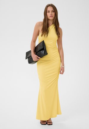 Femme debout portant une robe jaune longue asymétrique, tenant une grande pochette noire, avec des sandales noires et un bracelet au poignet.