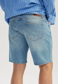 Shorts en denim bleu clair avec un ourlet effiloché, des poches arrière et un patch en cuir de marque, avec une coupe décontractée et une texture douce.