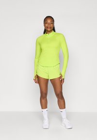 Top deportivo de color verde neón con cuello de cremallera y mangas largas, combinado con pantalones cortos a juego. Zapatillas y calcetines blancos de Nike completan el conjunto.