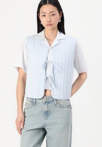 Lyseblå quiltet vest med krage og knytelukking, med vertikale striper. Brukt over en hvit skjorte og kombinert med lys denim jeans.