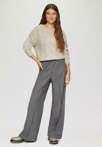 Beige Kabelstrickpullover mit entspannter Passform, kombiniert mit grauen, weit geschnittenen Hosen. Das Model trägt klobige Schuhe mit dunkler Sohle.