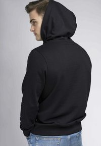 Sudadera con capucha negra y un acabado texturizado. Presenta un bolsillo canguro delantero y puños de canalé, combinada con jeans de color azul claro.