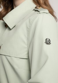 Lichtgroene jas met een kraag, knoopdetails en een logo patch op de mouw. Gladde textuur met een ruime pasvorm en zakken.