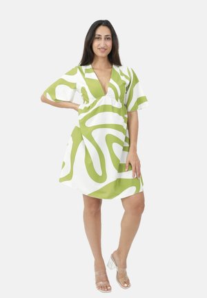 Mujer de pie con la mano en la cadera, vistiendo un vestido blanco con audaces patrones abstractos verdes y sandalias con tacón transparente sobre fondo blanco.