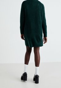 Una persona con la pelle scura indossa un vestito a maglia verde, calze bianche e scarpe con plateau nere, rivolta di spalle alla fotocamera.