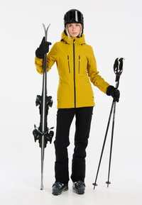 Giacca da sci gialla, pantaloni da sci neri, tiene gli sci e gli attacchi. La giacca presenta cerniere e un cappuccio. Stivali da neve visibili, casco e occhiali da sole indossati.