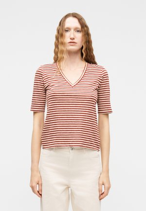 Woman with long wavy hair wearing a red and white striped V-neck short-sleeve top and white pants, standing against a plain background.