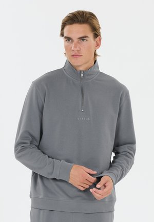 Jonge man die een grijze sweater met een kwartrits draagt, met "VIRTUS" gedrukt op de borst, die met beide handen de mouw aanpast.