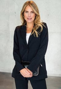 Zwarte op maat gemaakte blazer met twee knopen, gedragen over een witte blouse. Accessoires omvatten een gestructureerde zwarte clutch met zilveren studs. Eenvoudige achtergrond.