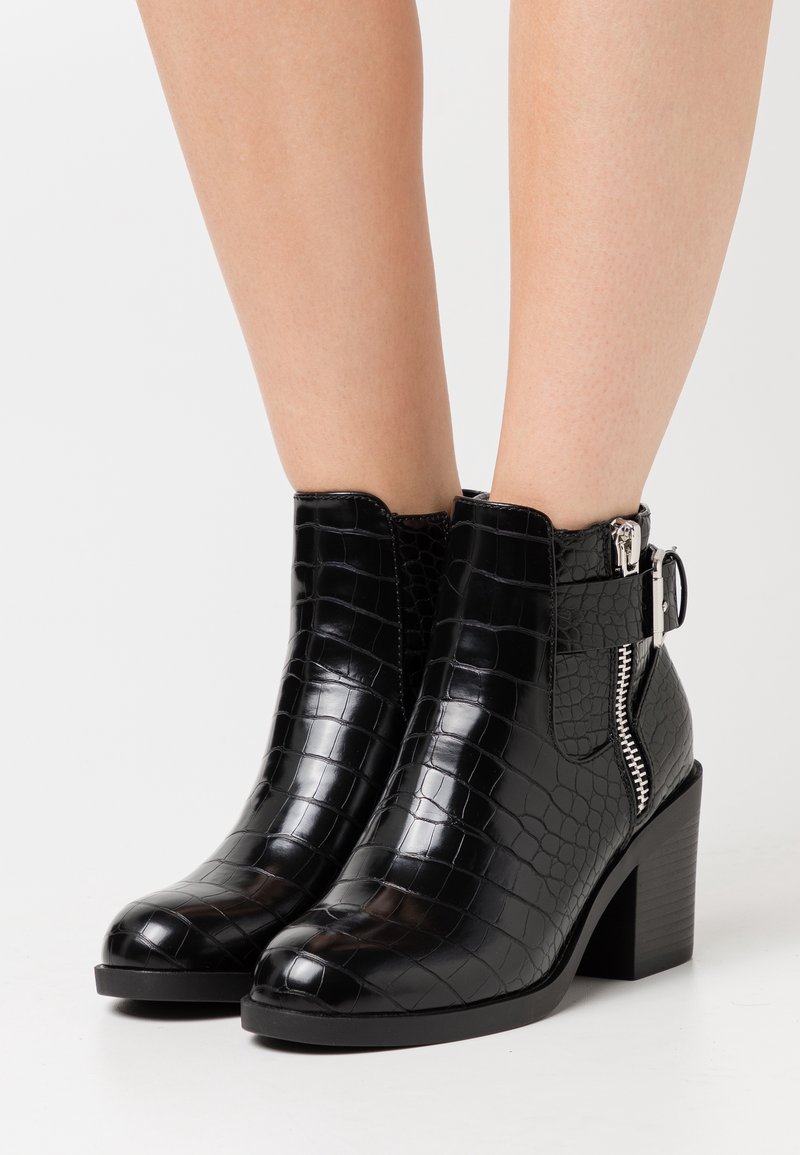 Pieds chaussés de bottines noires en cuir embossé effet croco, avec talons blocs, fermeture éclair latérale et détail de bride à boucle sur fond blanc.