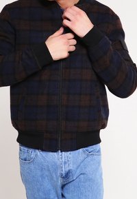 Henkilö, jolla on päällään tumma ruudullinen vetoketjupalttina ja vaaleansiniset farkut, kiinnittää takin vetoketjua kummallakin kädellä.