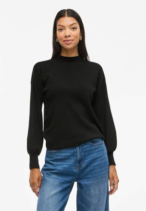 Pull en maille noire avec un col montant et de longues manches bouffantes. Le tissu semble doux, associé à un jean droit bleu clair.