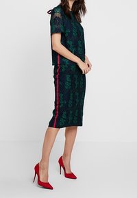 Mönstrad spetsblus i mörkgrön färg med blommönster, matchad med en åtsittande mörkgrön pennkjol med röda sidostråk och höga röda klackar.