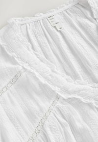 Blouse en tissu blanc texturé avec des détails en dentelle et un col à volants, avec une étiquette cousue visible avec du texte.