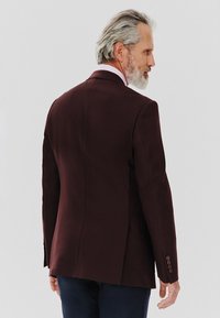 Burgundowa, dopasowana marynarka o strukturalnym kroju, z pojedynczym rozcięciem i detalami w formie trzech guzików przy mankietach. Gładka faktura i minimalistyczny design.