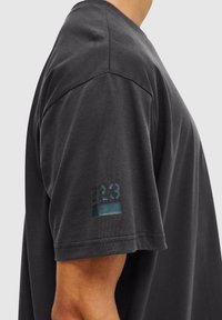 Zwarte katoenen t-shirt met een relaxte pasvorm, korte mouwen en een zachte textuur. Bevat een subtiele "223" grafiek op de linkermouw.